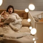 Uma foto muito acolhedora, com iluminação quente (amarelada), mostrando um pai ou mãe sentado numa poltrona a ler um livro para o bebé, ou a dar o biberão num ambiente calmo.